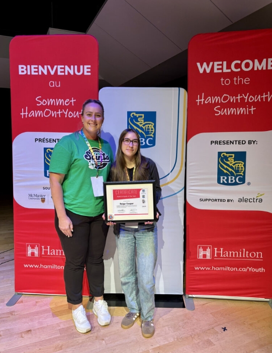 Teacher and student hold HamontYouth of the Year award frame infront of HamontYouth banners